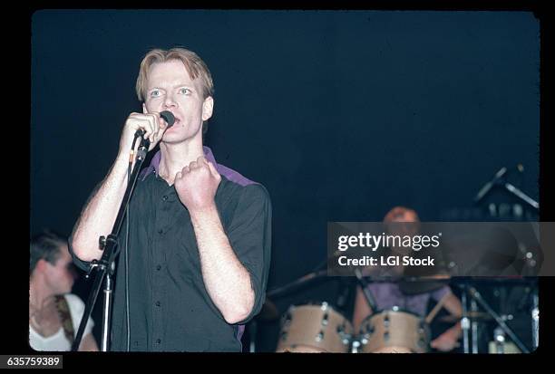 Picture shows writer/poet/musician, Jim Carrol, performing on stage. He is speaking into a microphone wearing a black button-down. There is s drummer...