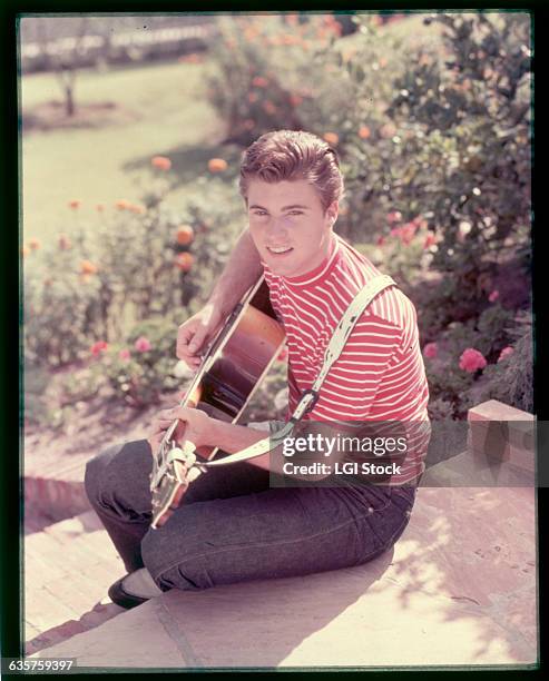 Picture shows teen idol/singer, Ricky Nelson, seated on a step outside playing the guitar. He is looking at the camera and wearing blue jeans and a...