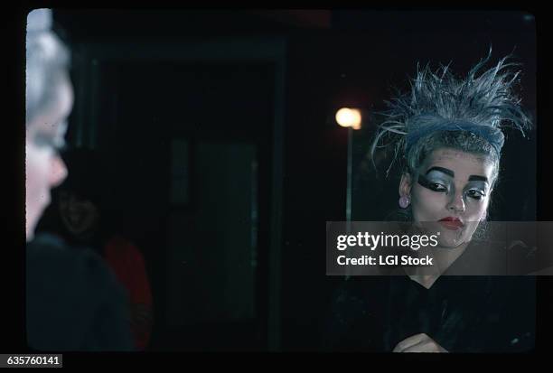 Singer Nina Hagen waist-up, reflected in a mirror. She is shown with dyed blue hair and heavy make-up. Photograph, 1980.