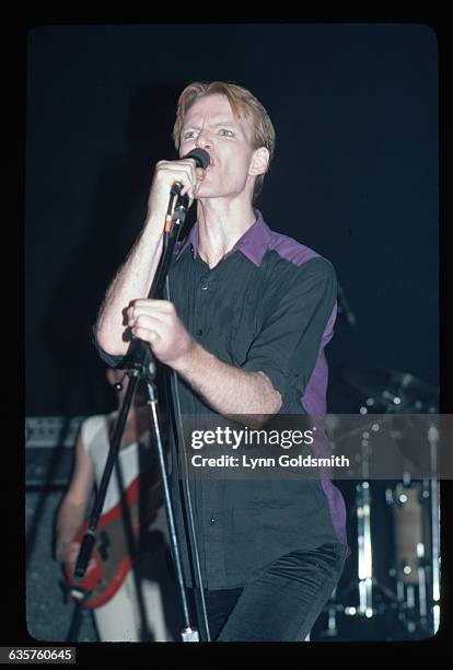 Picture shows writer/poet/singer, Jim Carrol, performing in concert. He is singing into a microphone, holding on to it with both hands. He is wearing...