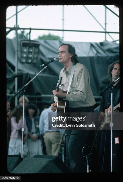 Singer/songwriter James Taylor is shown on stage playing guitar and singing into a microphone in concert.