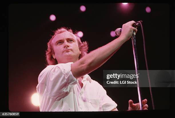 Picture shows singer, Phil Collins, holding a microphone on stage during a concert. He is wearing a white button-down with the sleeves rolled up to...