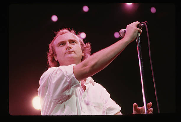 Picture shows singer, Phil Collins, holding a microphone on stage during a concert. He is wearing a white button-down with the sleeves rolled up to...