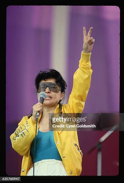 Yoko Ono performing. She is shown waist-up, wearing sunglasses and a yellow jacket. Photograph, 1987.