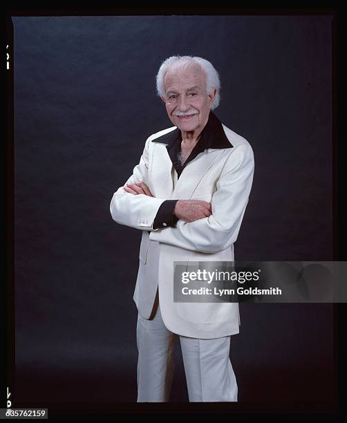 Picture shows conductor for the Boston Pops Orchestra, Arthur Fiedler, posing in a white suit with a black, wide collar shirt and his arms crossed in...