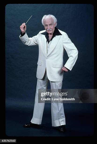 Picture shows conductor of the Boston Pops Orchestra, Arthur Fiedler, wearing a white suit and black, wide collar shirt. He is standing in front of a...