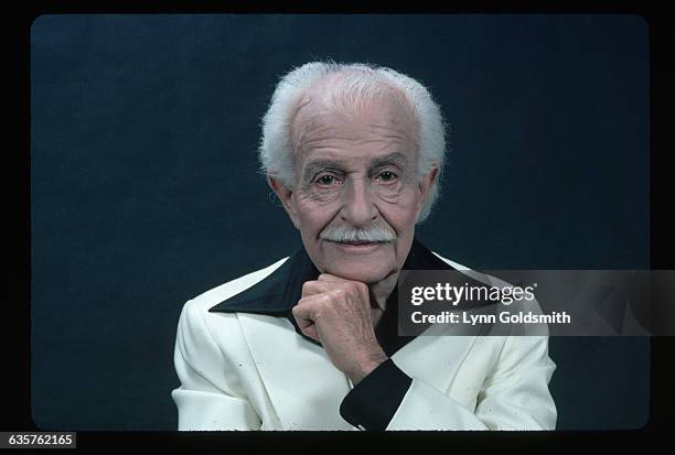 Portrait of Boston Pops Orchestra, Arthur Fiedler. He is dressed in a white suit and a black, wide collar shirt. His hand is placed on his chin and...
