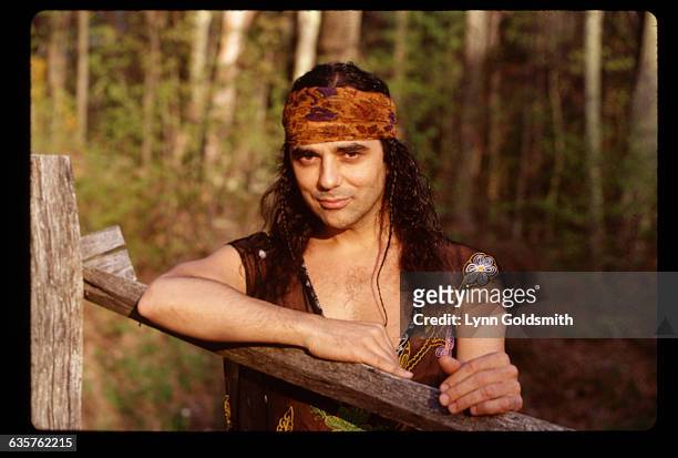 Producer and musician Daniel Lanois is shown outdoors, leaning on a wooden fence. He wears a bandana and a vest over his shirtless torso.