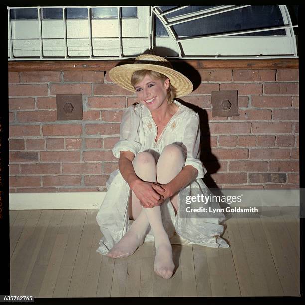 Singer Marianne Faithfull sits up against a wall with her knees bent to her chest. She wears a large straw hat. Undated photograph.