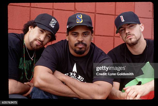 Left to right: B-Real, Sen Dog and DJ Muggs, of the group Cypress Hill, are shown seated in front of a red wall. All three wear baseball caps and...