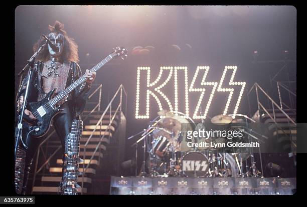 Musician Gene Simmons of glam-rock band sings "Kiss" standing on stage during a concert. He is playing his bass guitar in his costume and make-up. In...