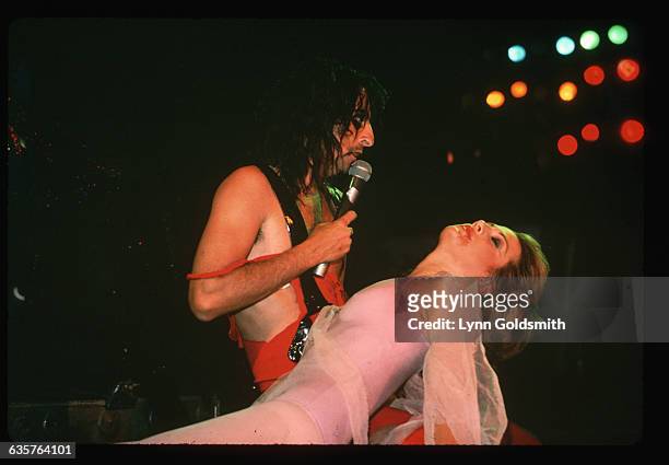 Hard rock musician Alice Cooper performing onstage, singing while holding his prone wife. Undated photograph.