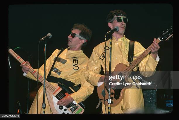 Two unidentified members of the band Devo are shown on stage, playing their guitars in concert. They wear yellow jumpsuits with Devo imprinted on...