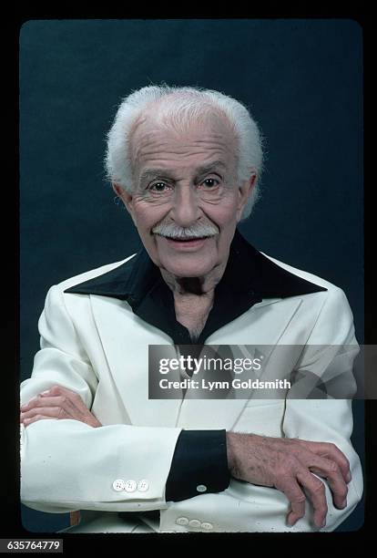 Classical music conductor Arthur Fiedler is shown in a studio portrait, arms folded. He wears a white jacket over a black shirt.