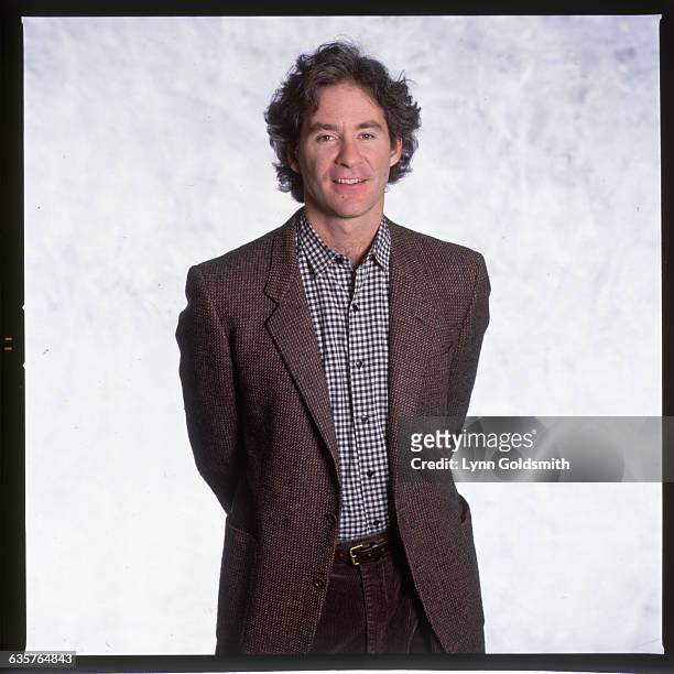 Picture shows actor, Kevin Kline, standing in a tweed jacket and check shirt with his hands behind his back.