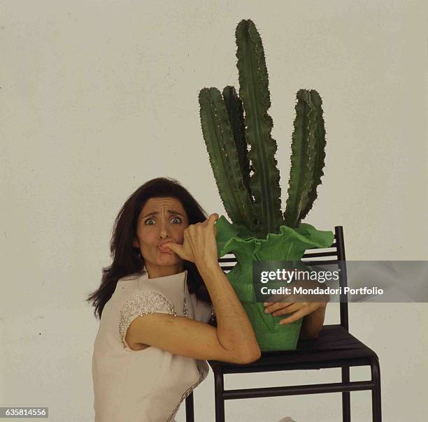 Italian comedian Anna Marchesini posing hugging a vase with a cactus resting on a high stool. Studio photo shooting. Italy, 1995