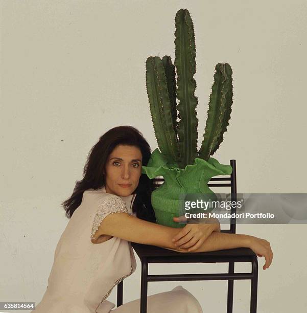 Italian comedian Anna Marchesini posing crouching and hugging a vase with a cactus resting on a high stool. Studio photo shooting. Italy, 1995