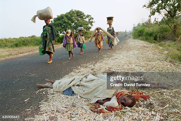 Refugees carrying bags walk near two bodies along the side of a road.