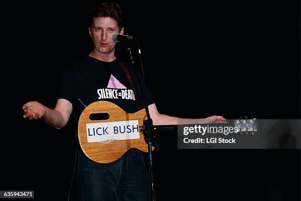 Billy Bragg is on stage showing off a guitar with a sticker that says, "Lick Bush," a joke about George Bush's run for the 1988 presidency.