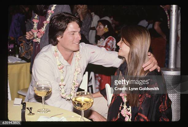 Christopher Reeve and Gae Exton gaze fondly at each other, wearing leis as they sit at a table over glasses of wine.