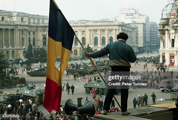 Man holds a Romanian flag with the Communist symbol torn from its center on a balcony overlooking the tanks, soldiers, and citizens filling Palace...