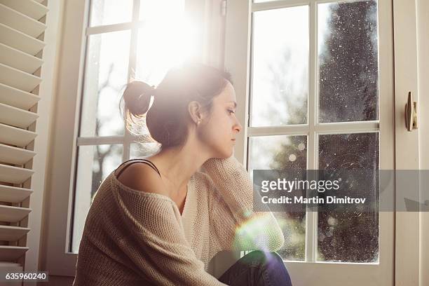 unglückliche hausfrau sitzt in der nähe des fensters - depression sadness stock-fotos und bilder