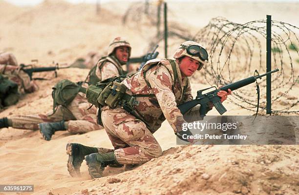 Marines crouch behind a barrier of barbed wire. They are training in a Saudi Arabian desert just before the onset of the Gulf War.