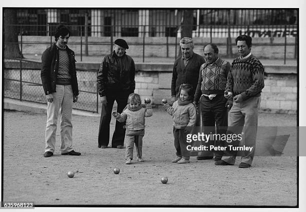 Elderly and middle-aged men watch two young girls play boules.