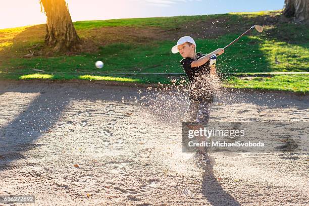 boy golfer hitting out a sand bunker - golf bunker stock-fotos und bilder