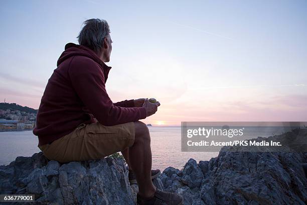 man relaxes on rocky bluff, watches sunrise, sea - sunrise contemplation stock pictures, royalty-free photos & images