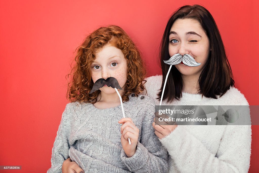 Woman and girl making faces with props mustaches, red wall.