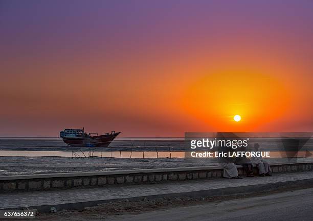 Men dressed in traditional dress, sitting on a bench in front of a sunset and a dhow boat on December 22, 2015 in Laft, Qeshm Island, Iran.