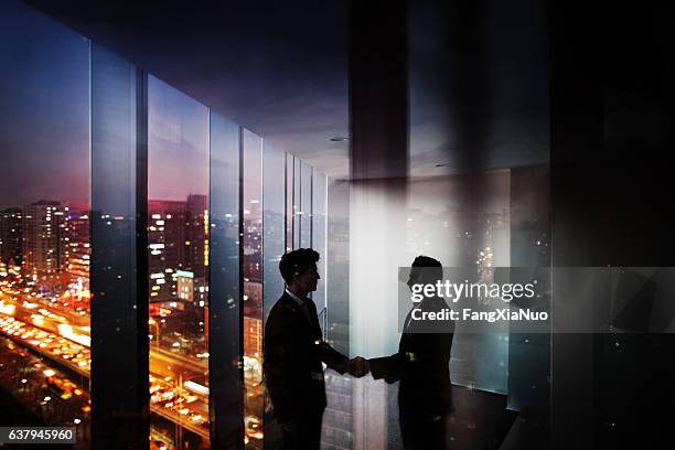 businessmen shaking hands in office at night with city view - secret handshake stock pictures, royalty-free photos & images