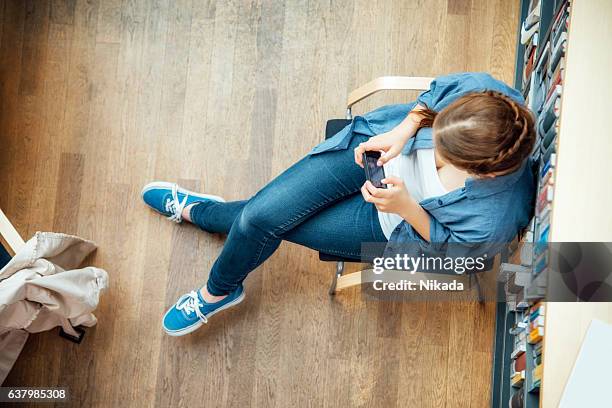 student using smart phone while sitting against bookshelf in classroom - woman elegant crossed legs stockfoto's en -beelden