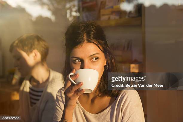woman drinking coffee in urban cafe while looking outside, sunny reflections in window. - kaffee trinken stock-fotos und bilder