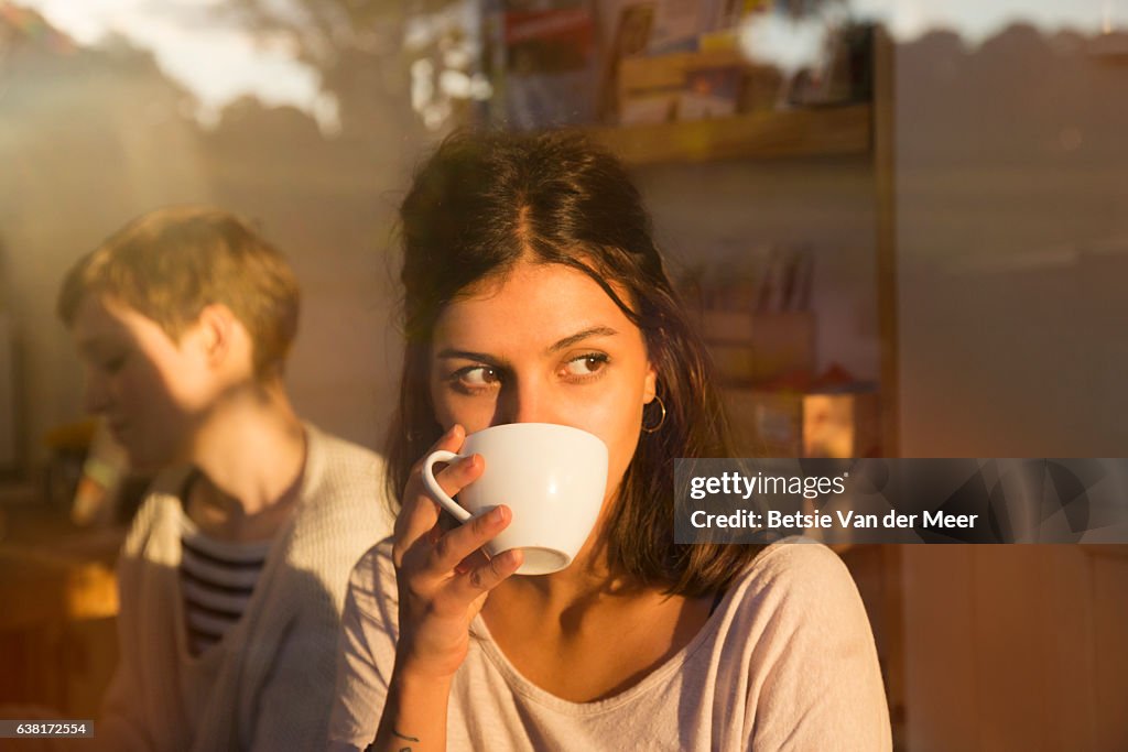 Woman drinking coffee in urban cafe while looking outside, sunny reflections in window.