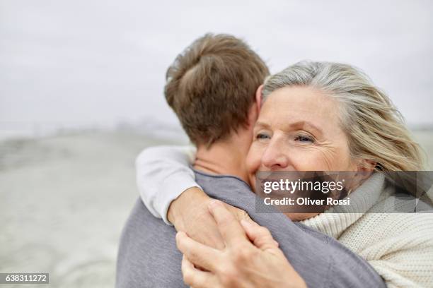 senior woman hugging adult son on the beach - senior man grey long hair stock-fotos und bilder