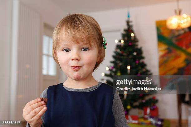 girl in front of christmas tree eating chocolate looking at camera - chocolate eating stock-fotos und bilder