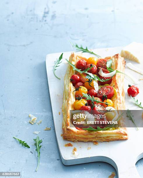 overhead view of tomato and parmesan puff pastry tart garnished with rocket on cutting board - savoury pie stock pictures, royalty-free photos & images