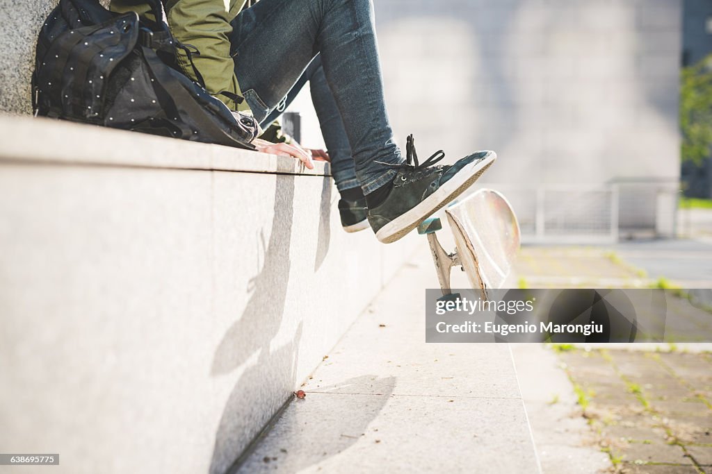 Waist down view of young male urban skate boarder sitting on wall flipping skateboard with feet