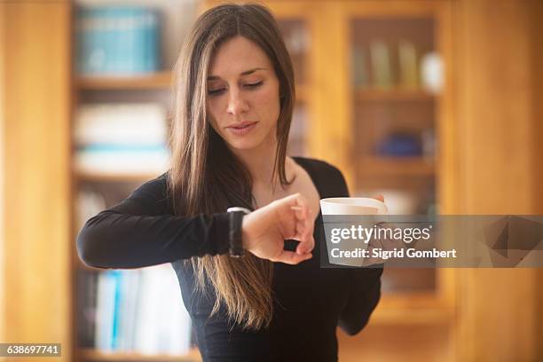 young woman at home, holding hot drink, looking at watch - important stock-fotos und bilder