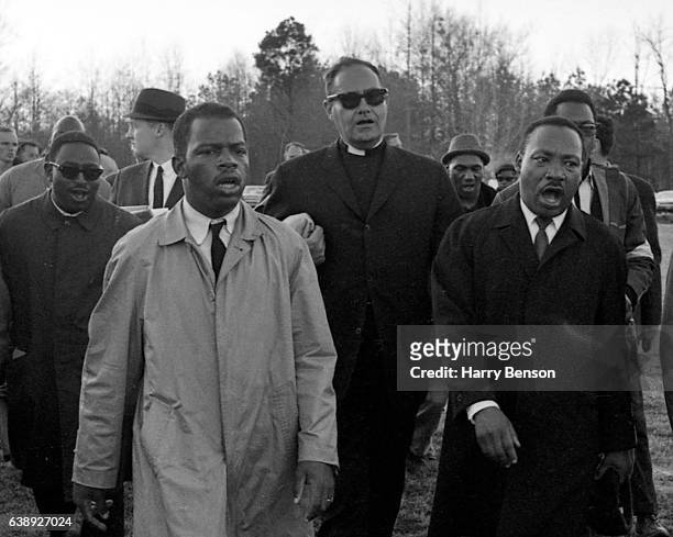 Dr. Martin Luther King, Jr. And Civil Rights Activist and U.S. Congressman John Lewis in light raincoat singing “We Shall Overcome” as they marched...