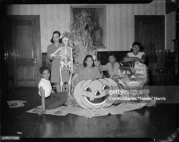 Five women, including Doris Harper second from right, posed with Halloween decorations including corn shock, jack-o-lantern, black cat, and skeleton...