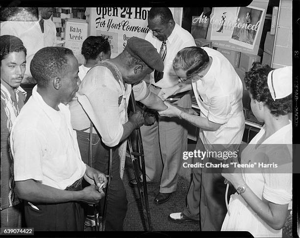 Dr W. A. Mason, Oliver Rake, and nurse Dora Lester Diuguid drawing blood of man on crutches, with Booker T. Williams on left, for blood testing...