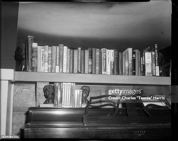 Rows of books including 'The Honey-Pod Tree' and 'The Langston Hughes Reader' held with portrait bookends on fireplace mantel and piano in front of...