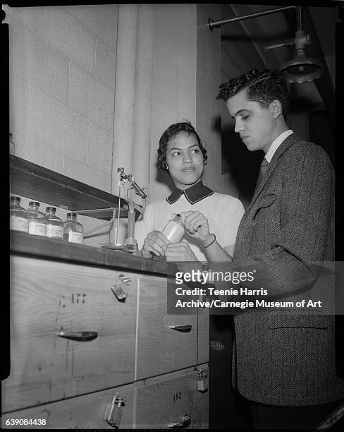 Young man and young woman standing at lab bench with drawers numbered 177 and 182, scale, and four glass bottles labeled 'Acetic...', 'Ammonium...',...