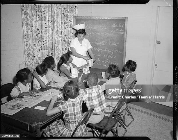 Eunice Cook wearing nursing uniform with cross on hat and circular brooch at collar, holding folded newspaper, standing at end of table with two boys...