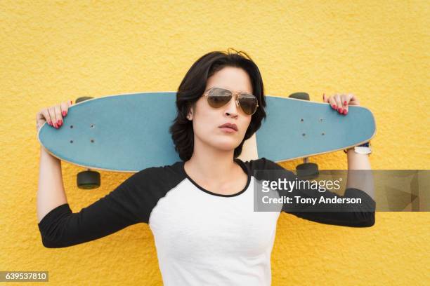 Young Hispanic woman standing near yellow wall with skateboard