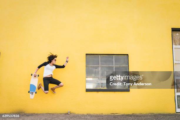 Young Hispanic woman jumping near yellow wall with skateboard