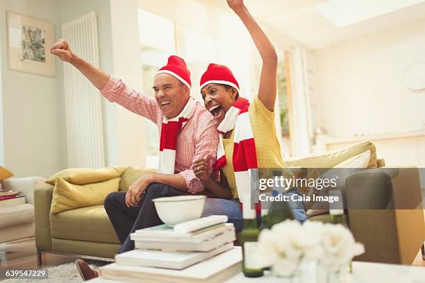 enthusiastic mature couple in matching hats and scarves cheering watching tv sports event - football wives and girlfriends - fotografias e filmes do acervo